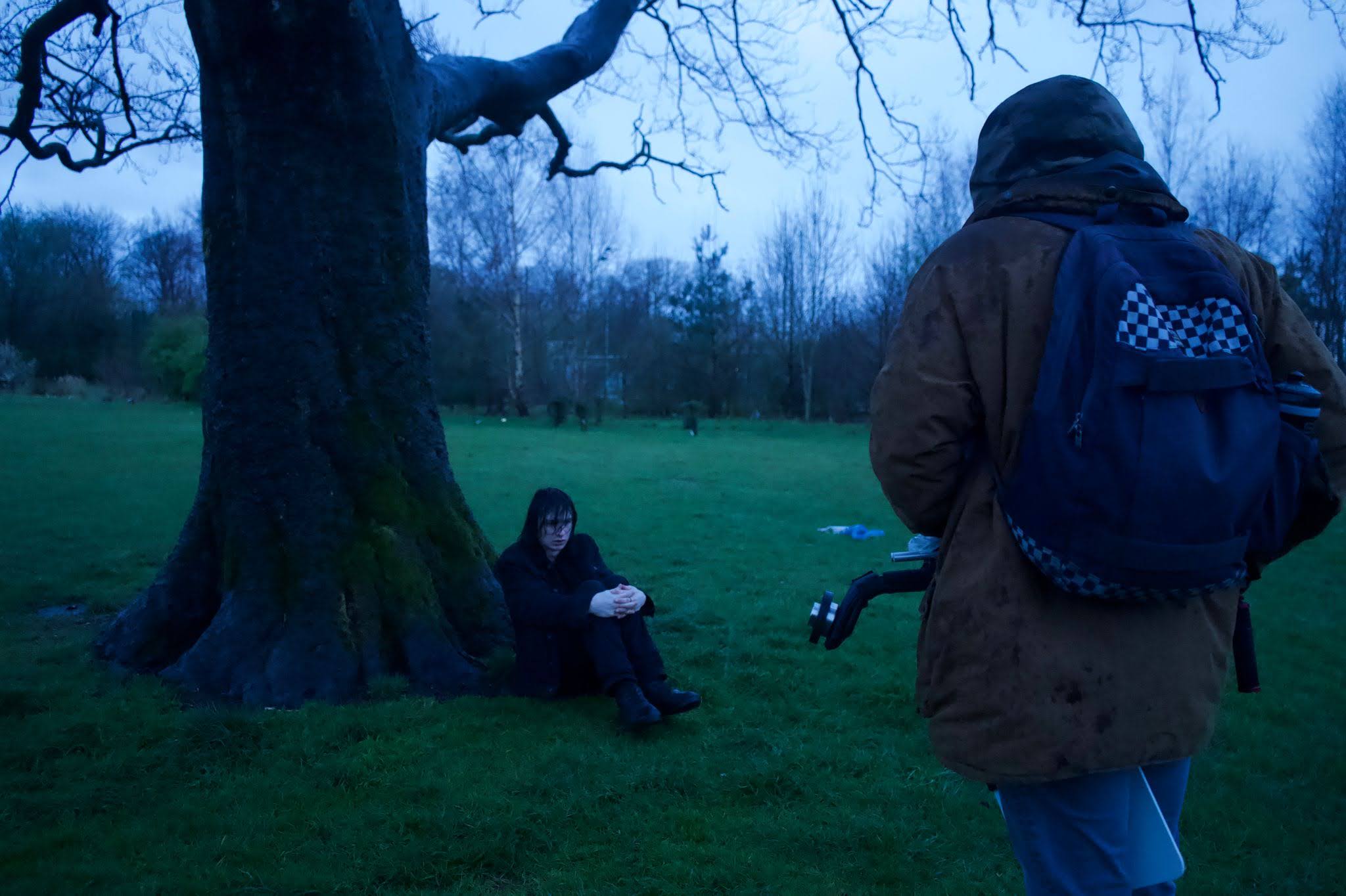 A man huddled under a tree, being filmed by crew.