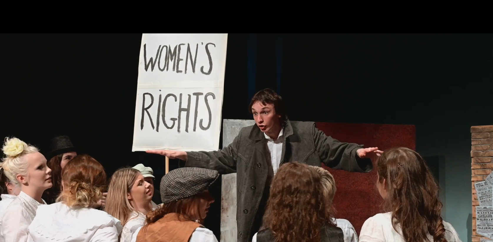A man giving an impassioned speech. A women's rights placard is in the background